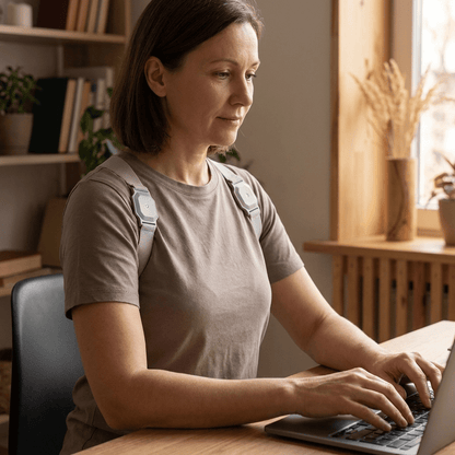 Woman sitting upright at a desk wearing the grey posture corrector over her shoulders while using a laptop